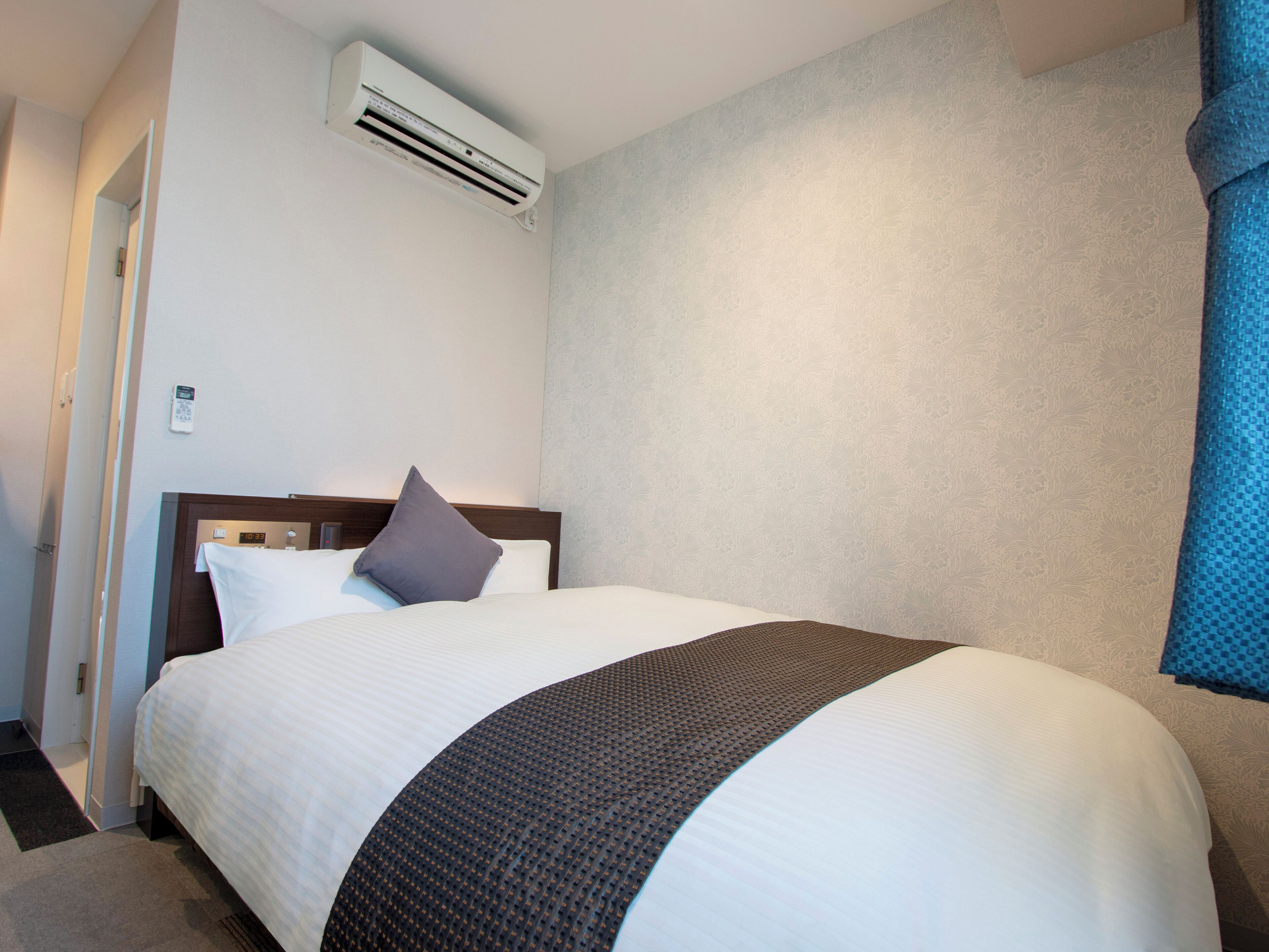 A hotel room with a bed, an air conditioning unit, a wall clock, and a door.