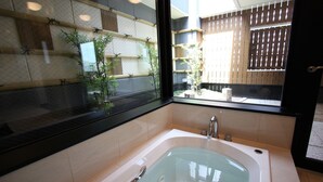 A modern bathroom with a large bathtub, a window with a view of a wooden fence and plants, and a tiled floor.