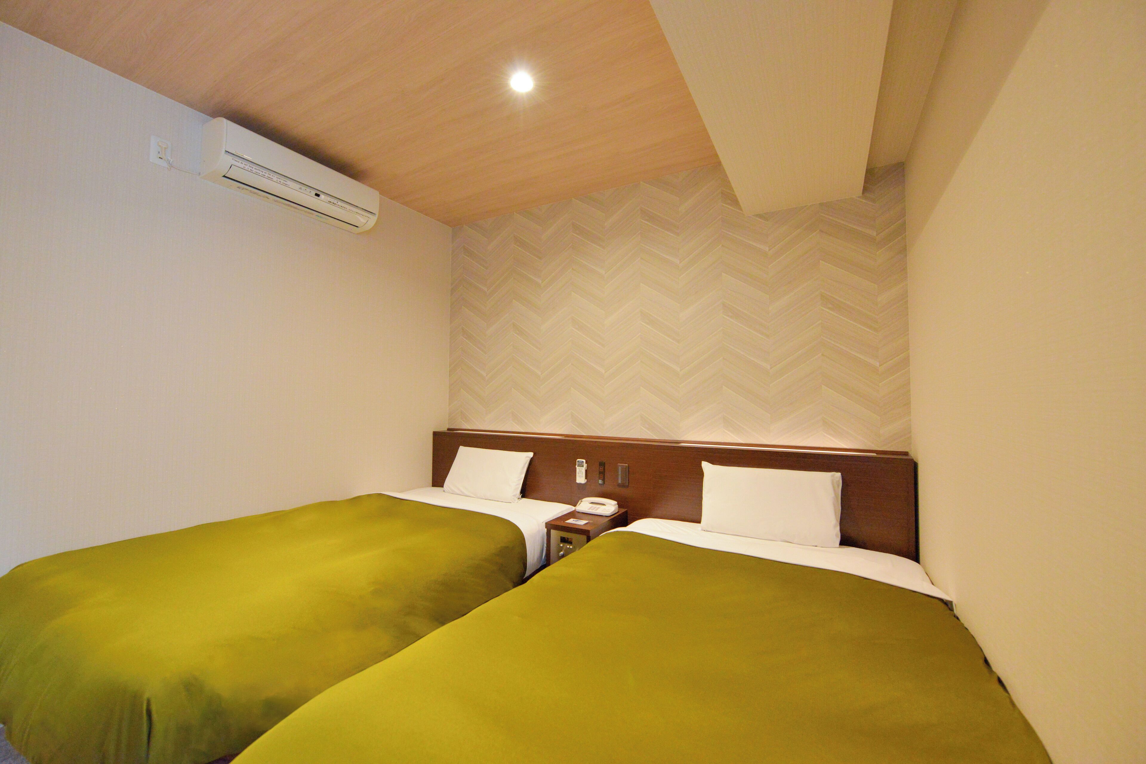 A hotel room with two single beds, a wooden headboard, a nightstand with a phone, and a wall-mounted air conditioner.