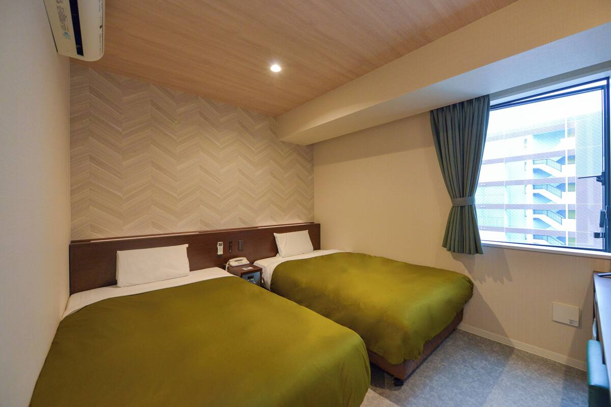 Habitación de hotel con dos camas, techo de madera, pared con diseño de espiga y ventana con cortinas, ofreciendo vista a un edificio cercano.