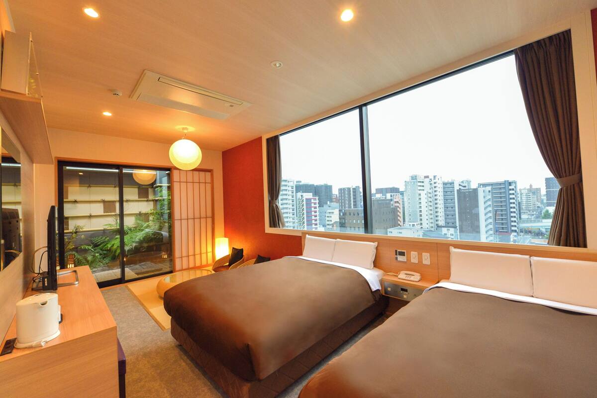 Habitación de hotel con dos camas, un escritorio, una silla, una ventana con vistas a la ciudad y un pequeño balcón con plantas.