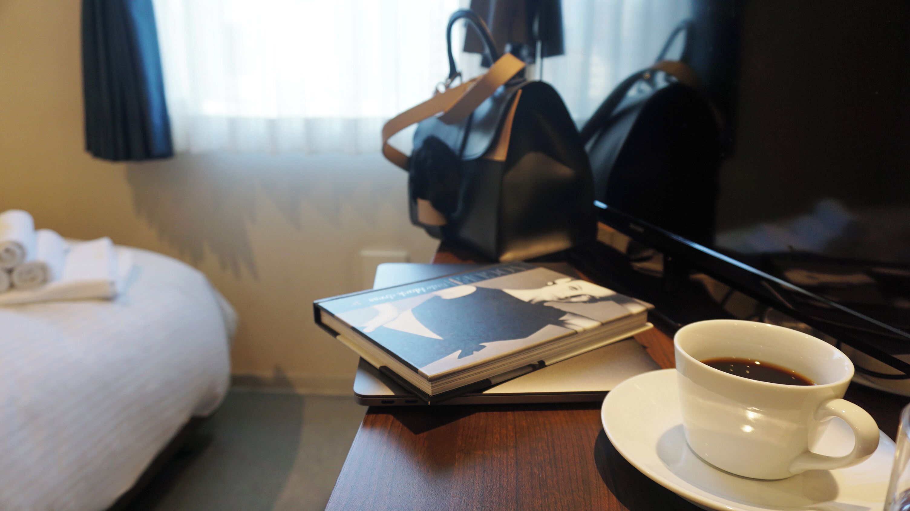 Habitación de hotel con una cama, una mesita de noche con un libro, una taza de café y un televisor.
