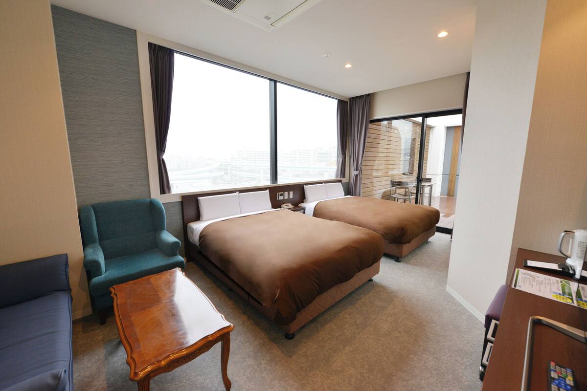 Habitación de hotel con dos camas, un sofá, una mesita y vistas a la ciudad a través de amplios ventanales.