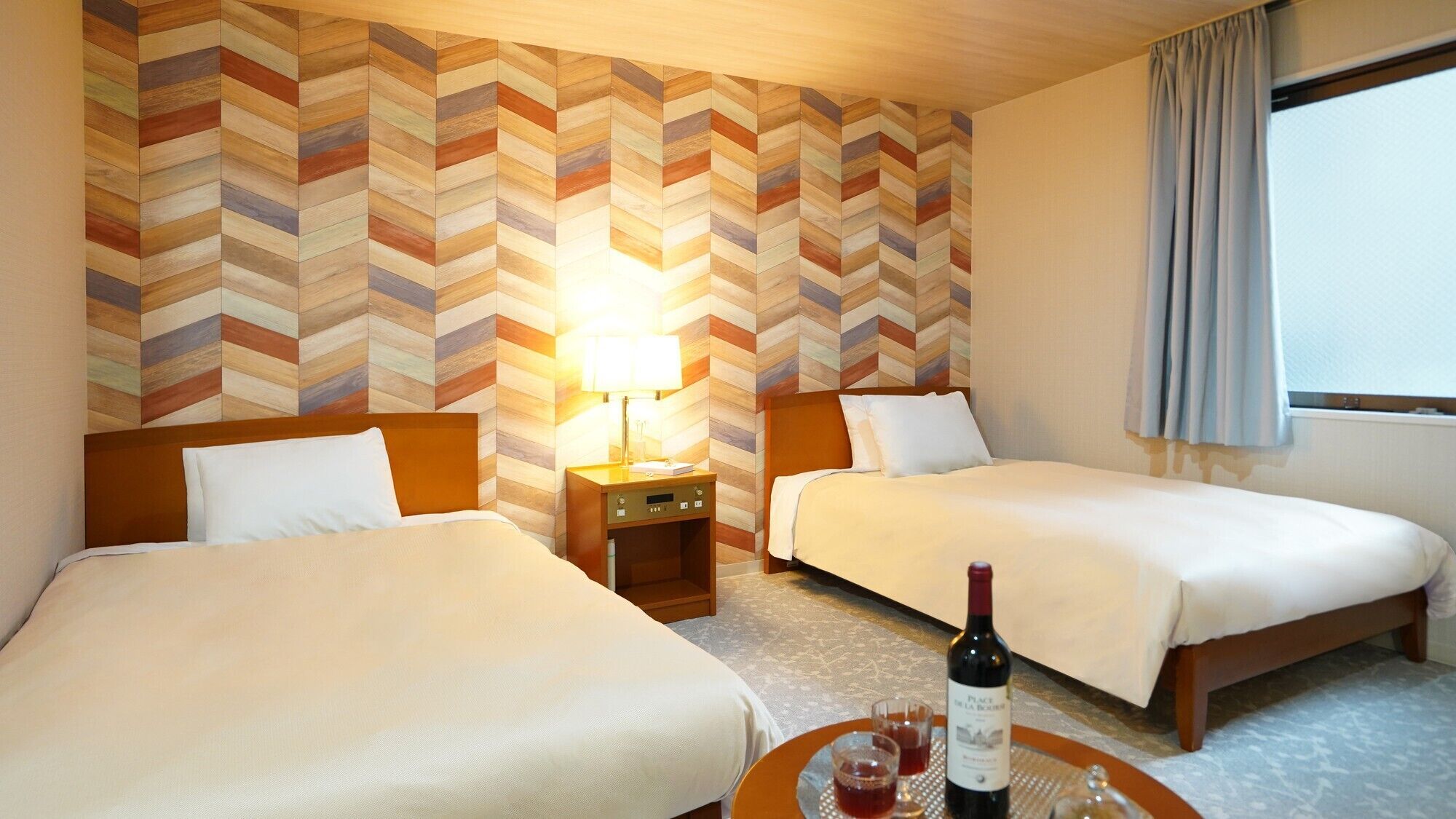 A hotel room with two beds, a nightstand, a lamp, a bottle of wine, and a view of the outside.