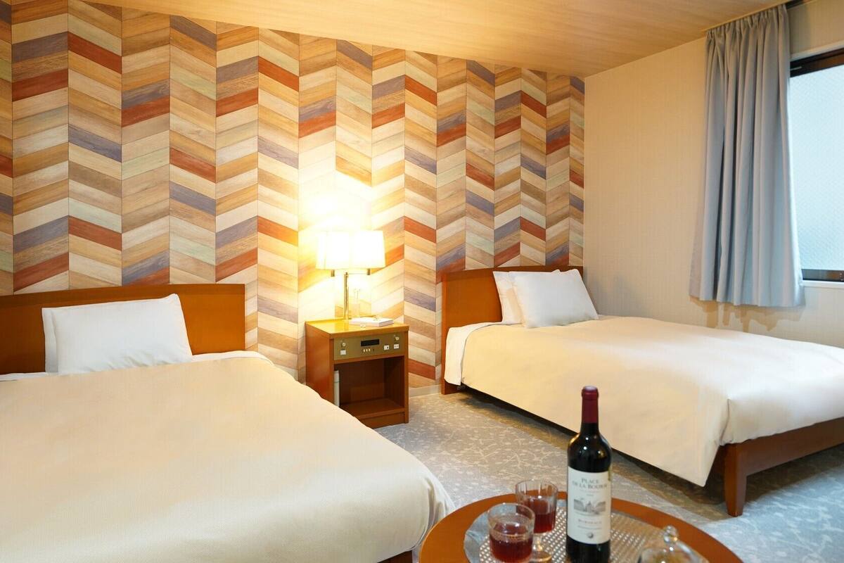 A hotel room with two beds, a nightstand, a lamp, a bottle of wine, and a view of the outside.