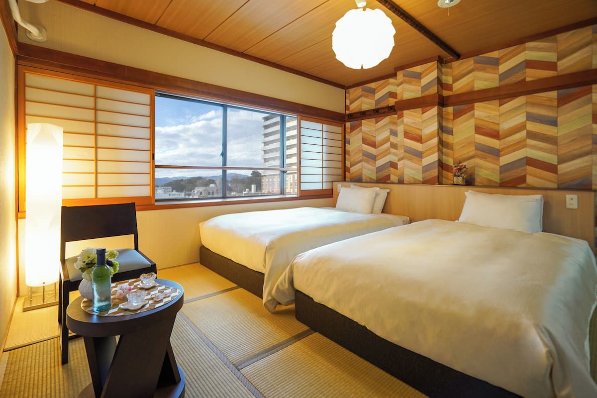 A traditional Japanese-style hotel room with tatami flooring, two beds, a window with a city view, and a decorative wall pattern.