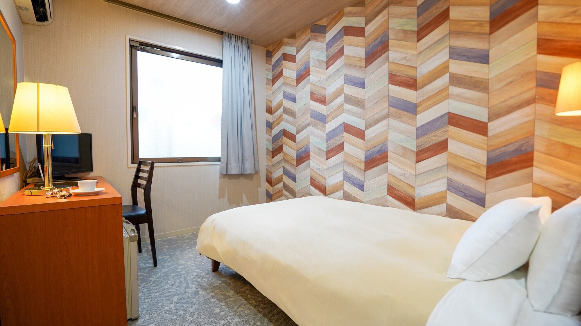 A hotel room with a bed, a desk with a lamp, a chair, a television, and a window with curtains.