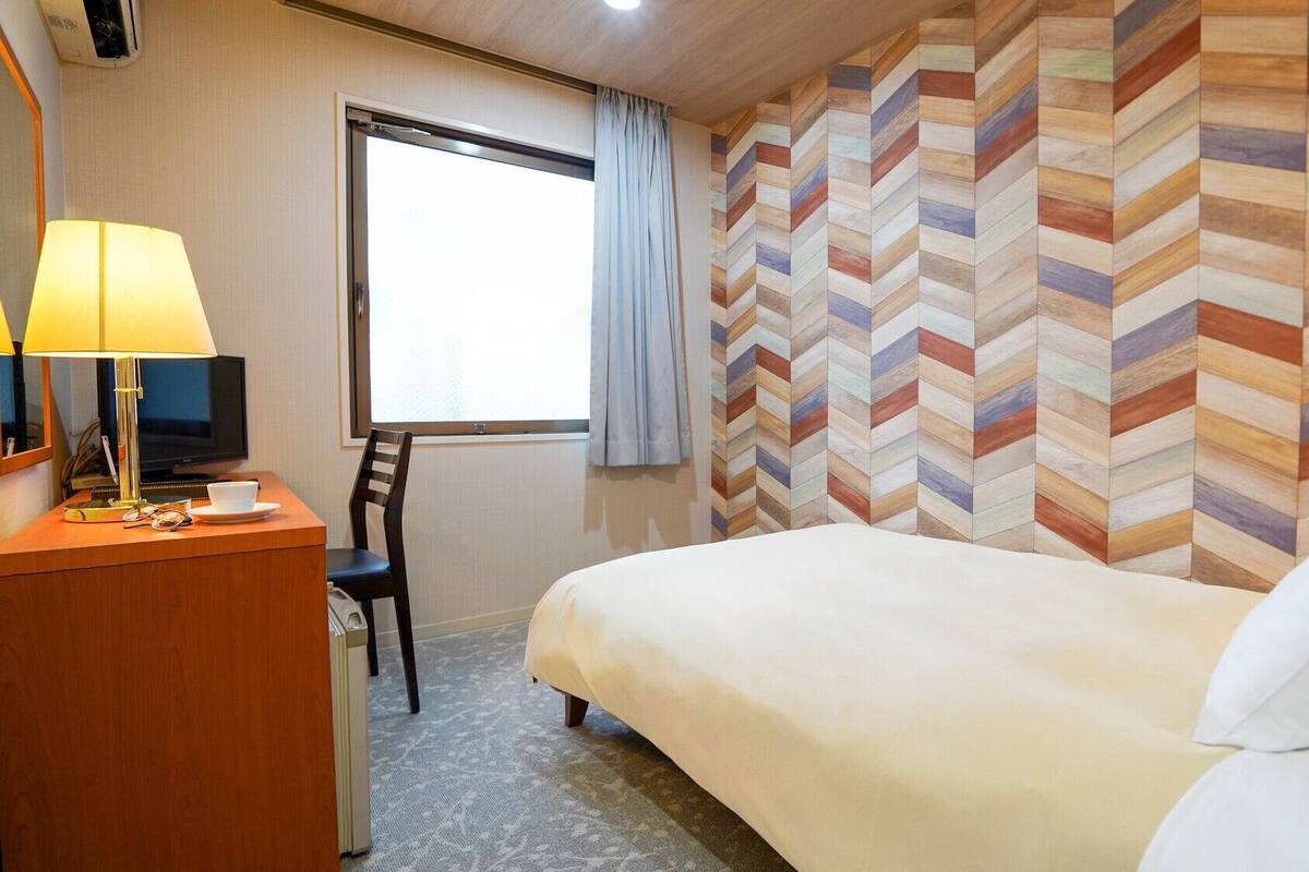 A hotel room with a bed, a desk with a lamp, a chair, a television, and a window with curtains.
