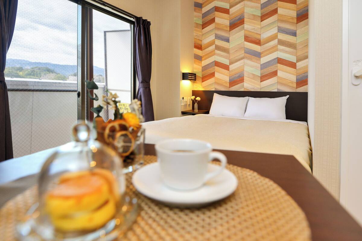A hotel room with a bed, a bedside table with a cup and pastries, a view of the outdoors, and a chevron-patterned wall.