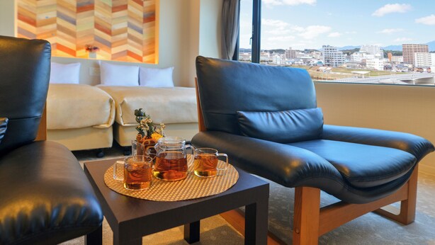Een moderne hotelkamer met uitzicht op de stad, een leren bank, een tafeltje met thee, en een decoratief kunstwerk aan de muur.