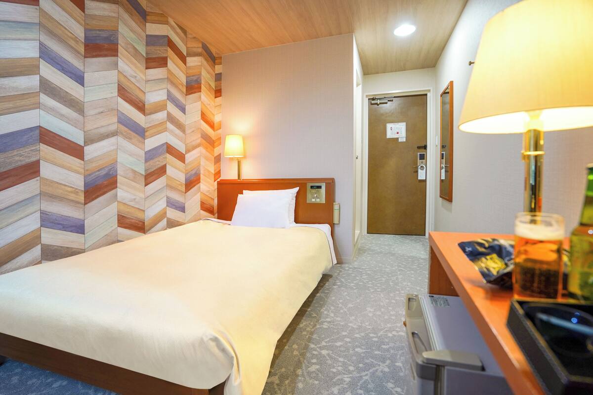 A hotel room with a bed, a desk with a lamp, and a door.