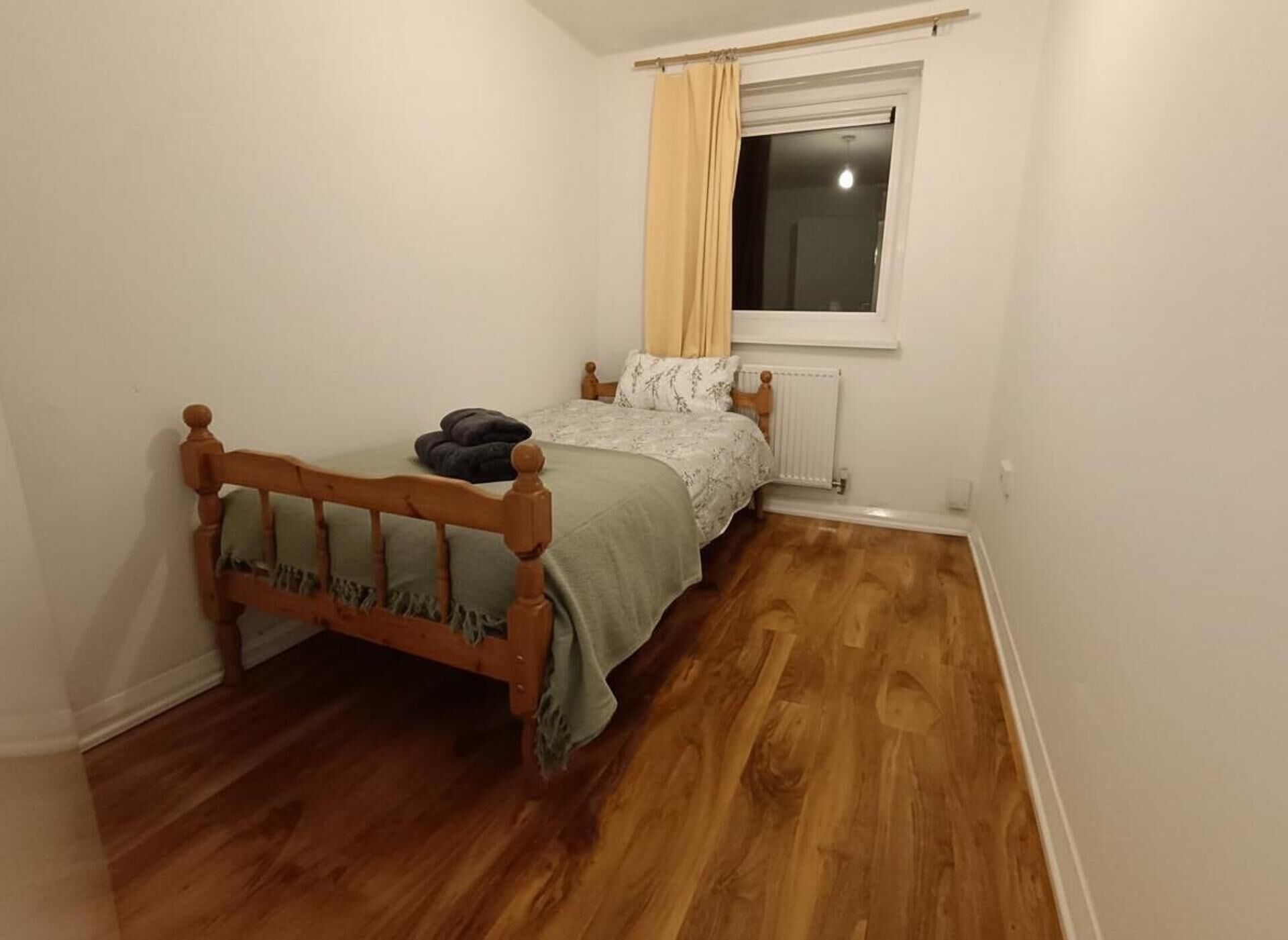 Une petite chambre avec un lit simple, une tête de lit en bois, un radiateur et une fenêtre avec des rideaux.