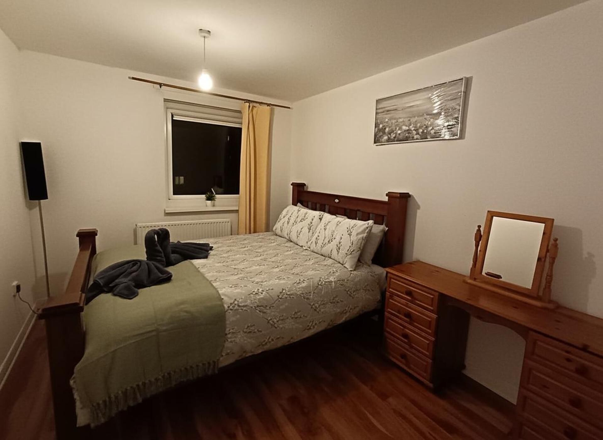 A bedroom with a wooden bed, a bedside table, a small dresser, a window with curtains, a radiator, and a wall-mounted picture.