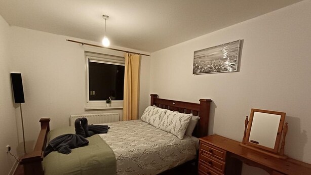 Une chambre à coucher avec un lit en bois, une table de chevet, une petite commode, une fenêtre avec des rideaux, un radiateur et un tableau accroché au mur.
