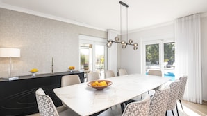 Un comedor moderno con una mesa de mármol, sillas estampadas, una isla negra con un cuenco de fruta y ventanas amplias con cortinas.