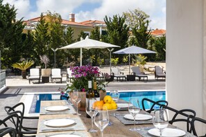 A poolside dining area with tables set for two, outdoor umbrellas, and a pool in the background. - Villa Alegria (Porches)