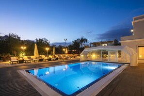 A swimming pool with lounge chairs and umbrellas, surrounded by modern buildings and palm trees. - Villa Alegria (Porches)