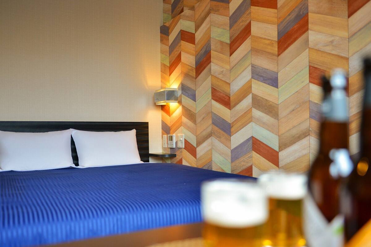 A hotel room with a bed, a wall with a herringbone pattern, a bedside table with a lamp, and a bottle of beer on the floor.