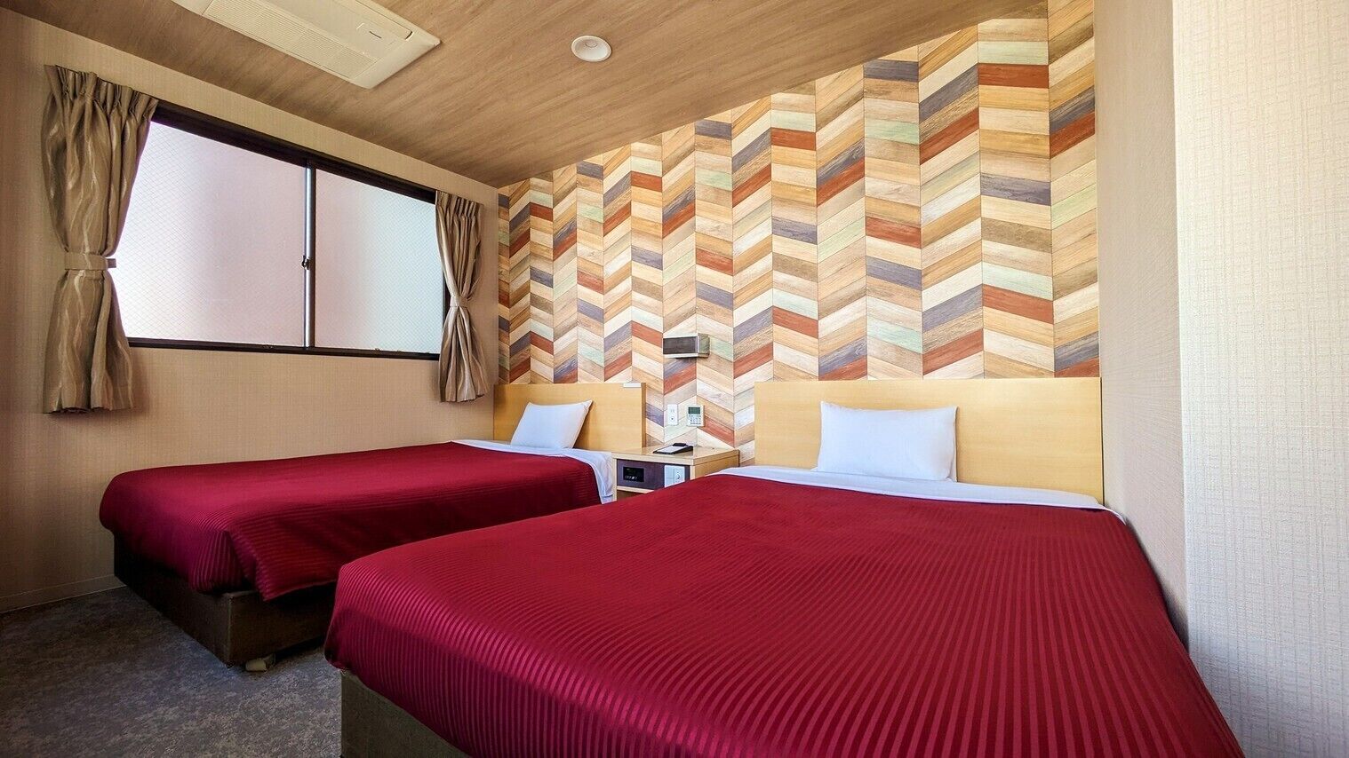 A hotel room with two beds, a wooden headboard, a herringbone-patterned wall, a window with curtains, and a ceiling light.