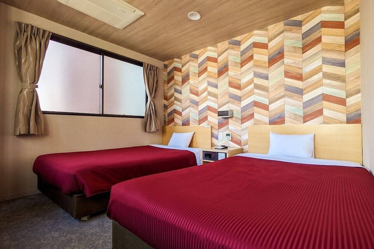 A hotel room with two beds, a wooden headboard, a herringbone-patterned wall, a window with curtains, and a ceiling light.