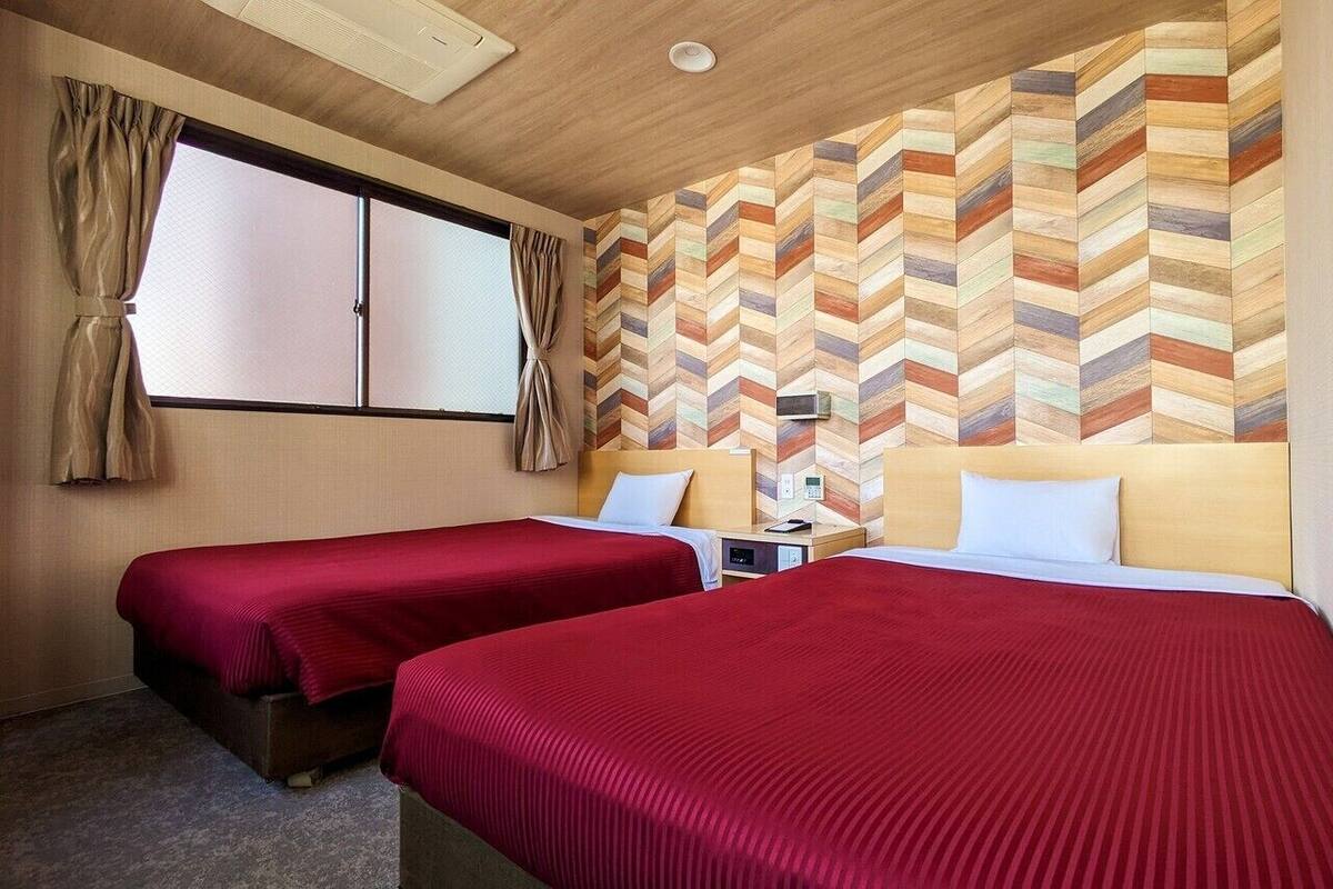 A hotel room with two beds, a wooden headboard, a herringbone-patterned wall, a window with curtains, and a ceiling light.