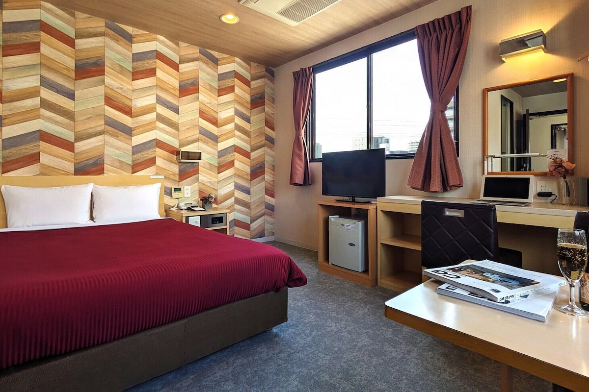 A hotel room with a large bed, a desk with a computer, a TV, and a window with curtains.