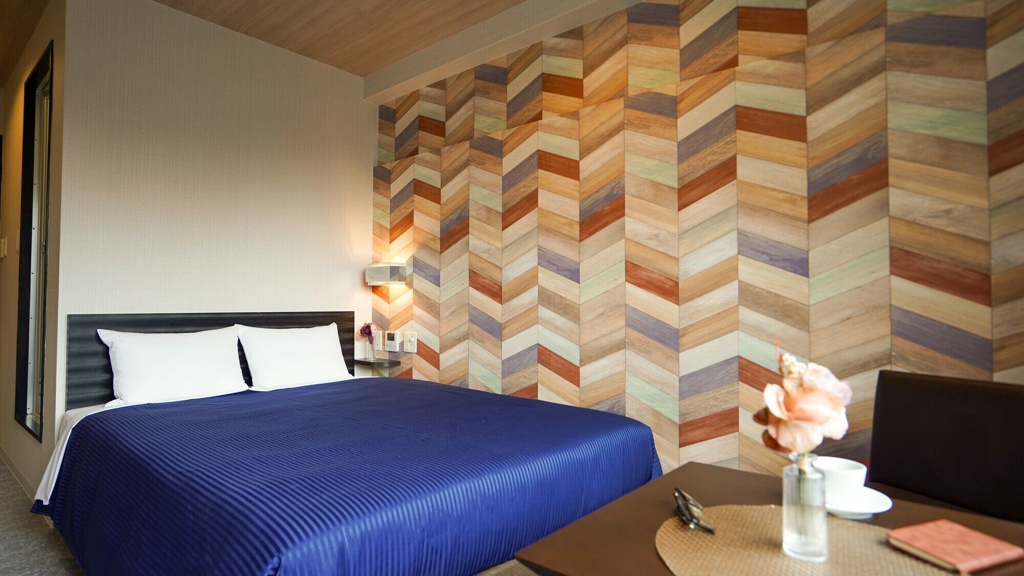 A hotel room with a bed, a nightstand, a wall with a chevron pattern, and a table with a vase of flowers.
