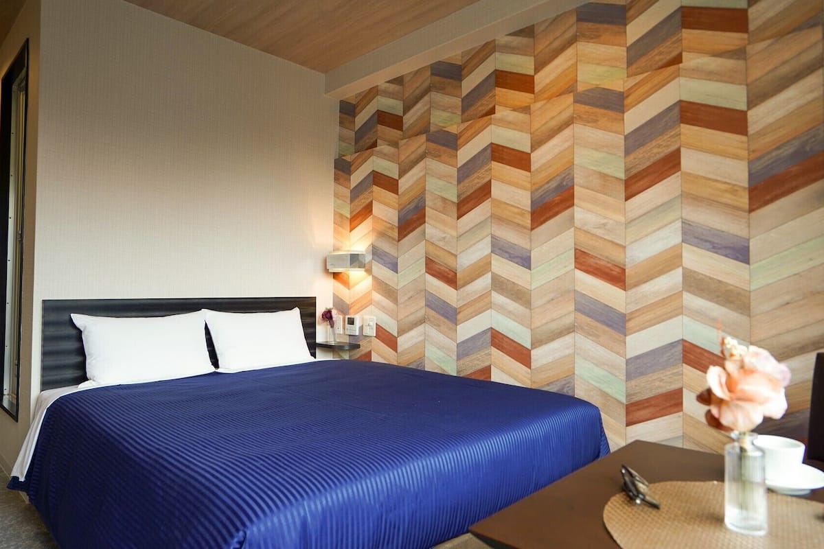 A hotel room with a bed, a nightstand, a wall with a chevron pattern, and a table with a vase of flowers.