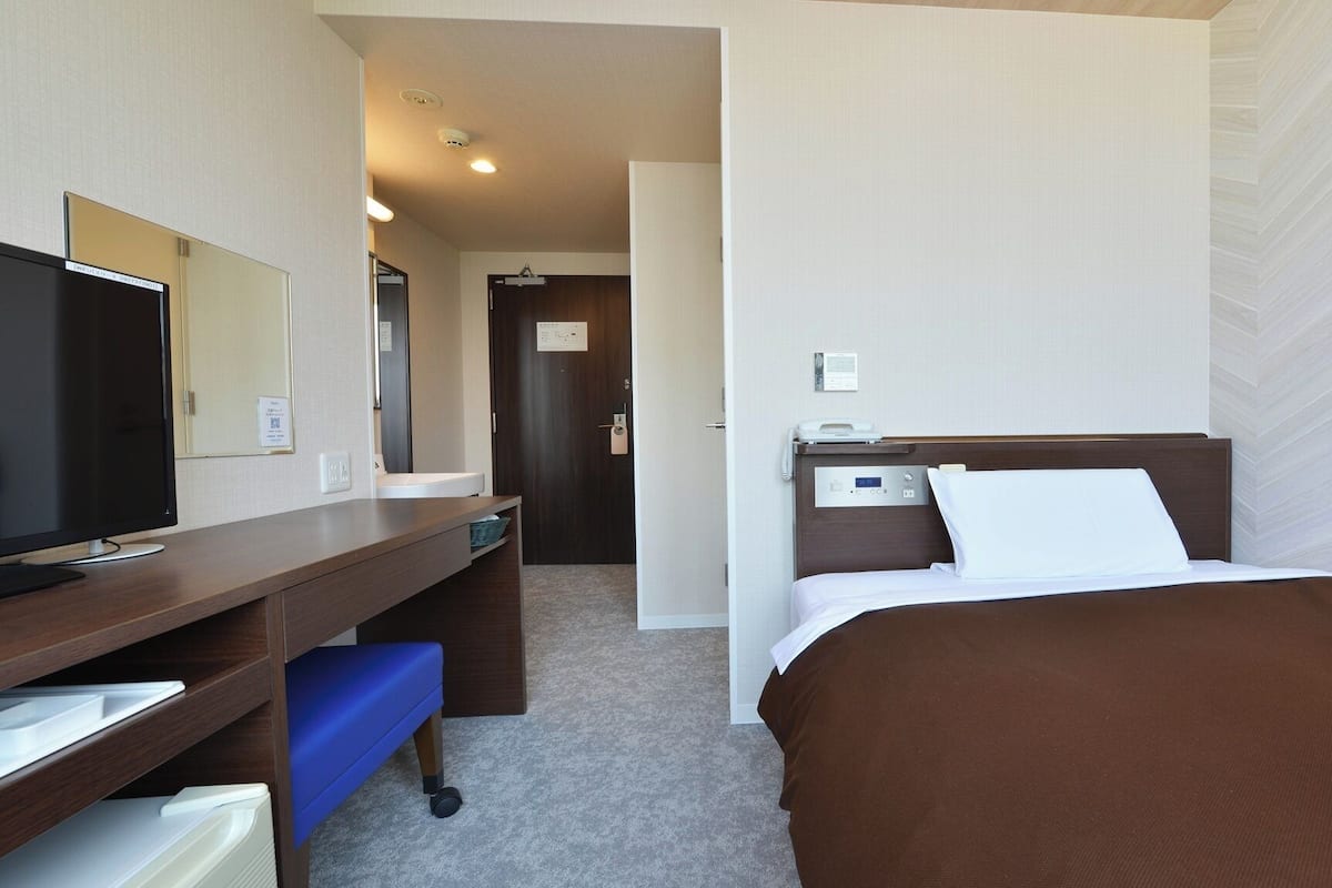 A hotel room with a bed, a desk with a computer, a TV, a bathroom, and a door.