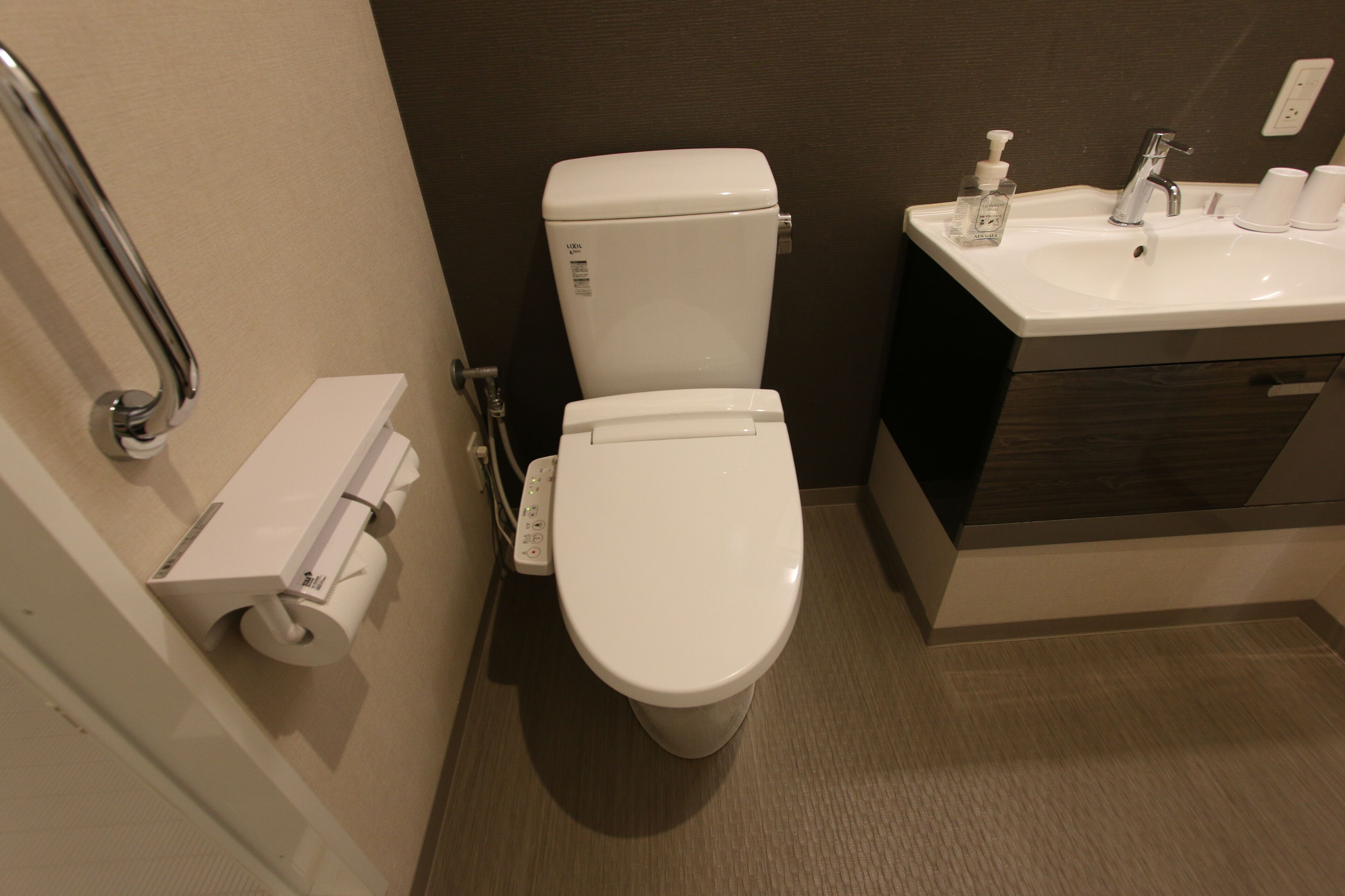 A modern bathroom with a toilet, a handrail, a sink with a soap dispenser, and a toilet paper holder.