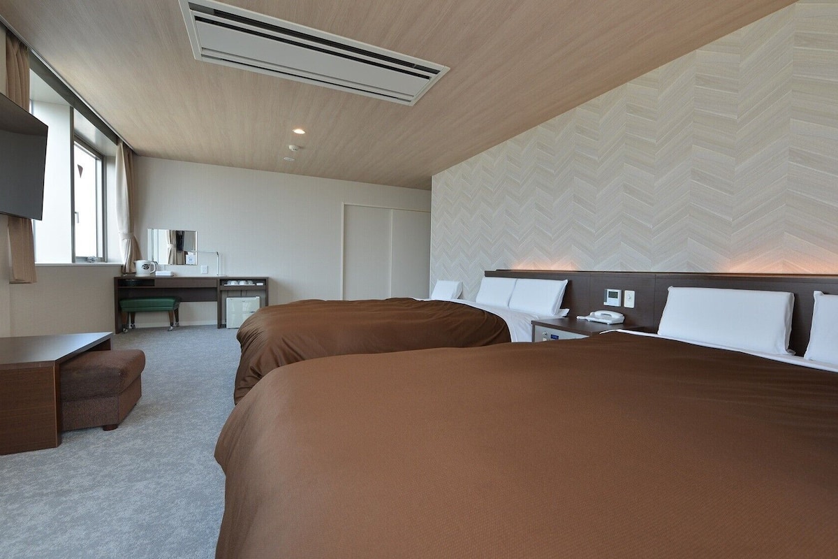A hotel room with two beds, a desk, a chair, a TV, and a large window.