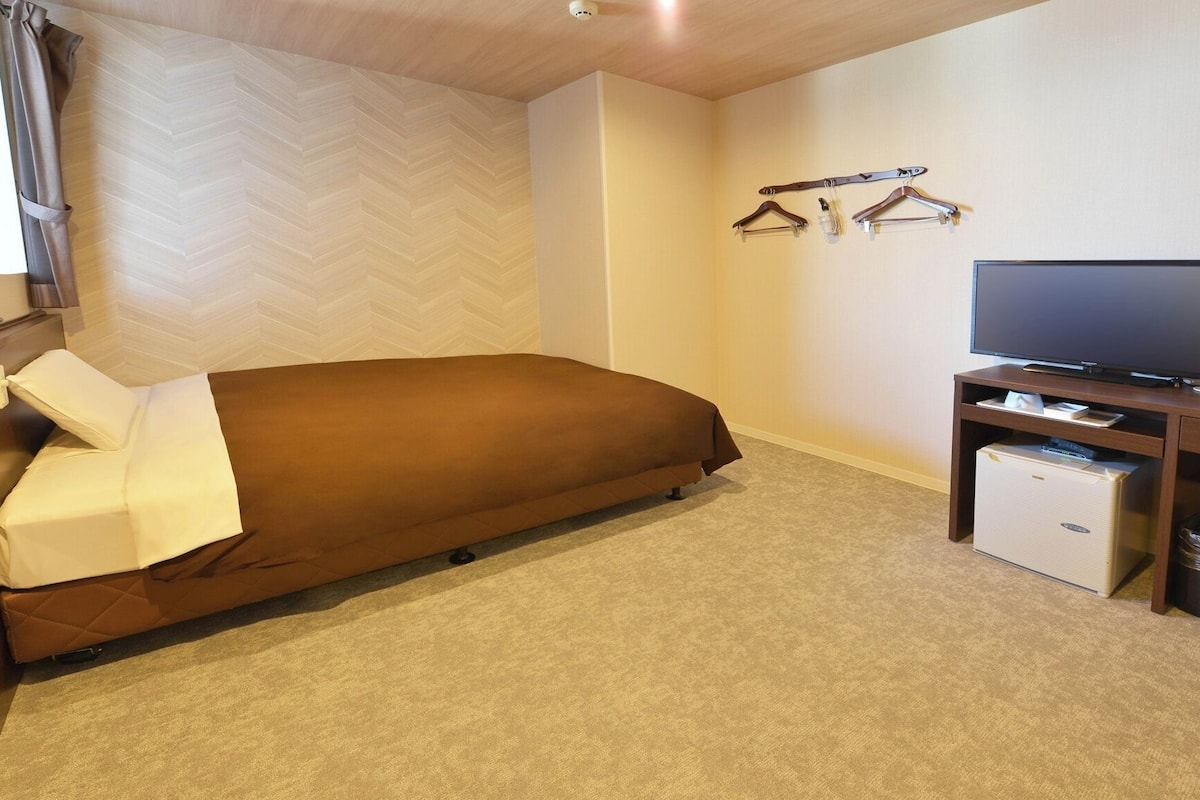 A single bed with a brown cover, a television on a wooden stand, and a small white appliance on the floor.