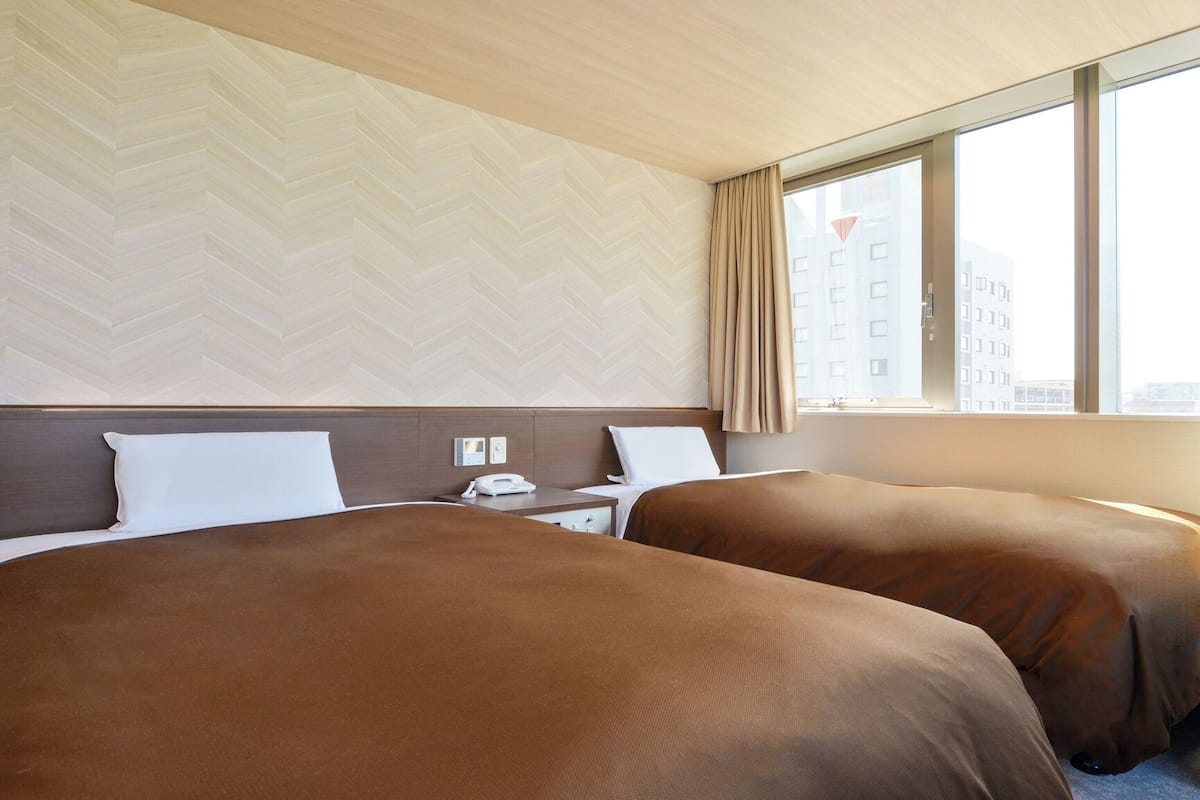 A hotel room with two beds, a wooden headboard, a nightstand, a telephone, and a window with a view of buildings.