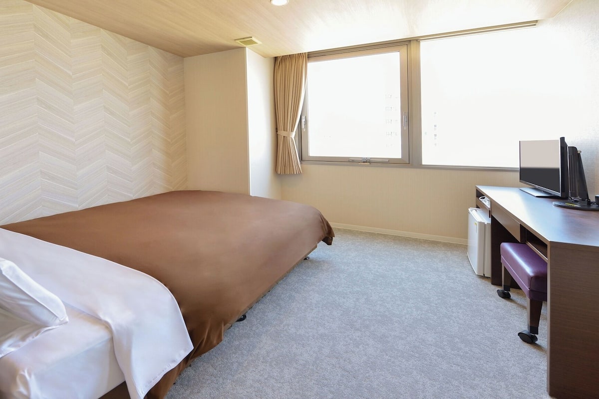 A hotel room with a bed, a desk with a computer, a chair, and a window with curtains.