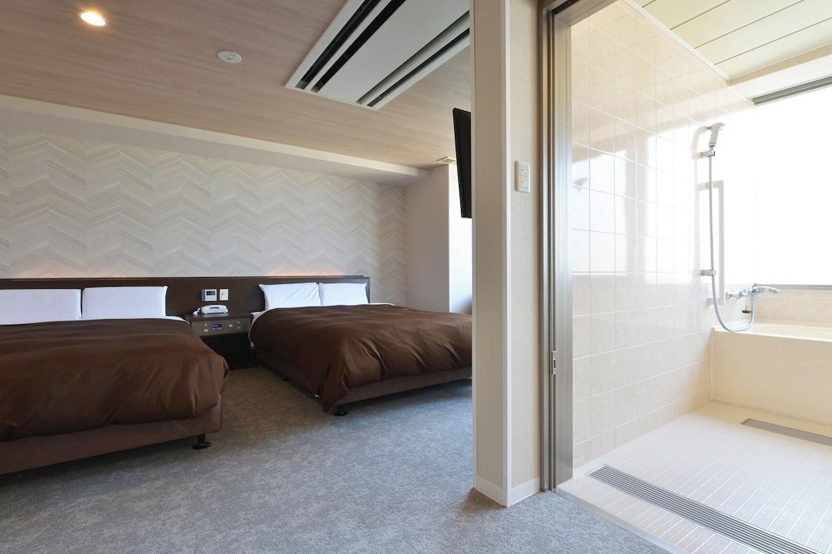 A hotel room with two beds, a bathroom with a bathtub, and a large window.