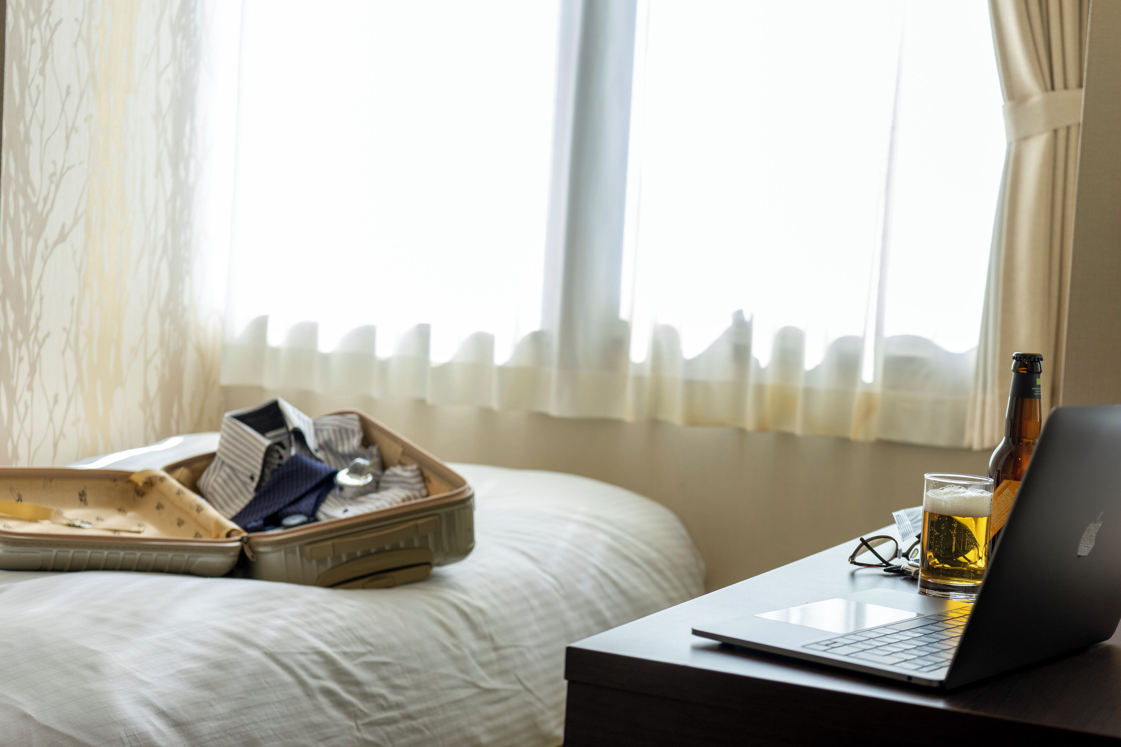 A hotel room with a bed, a suitcase, a laptop, a beer bottle, and a glass of beer on a nightstand.