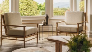 Dos sillones beiges de madera, una mesita redonda y un florero sobre una mesita auxiliar, todo en una habitación con ventanas amplias y vistas a zonas verdes.