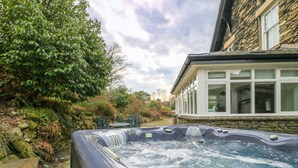 Un jacuzzi en un jardín con muros de piedra y una casa al fondo.