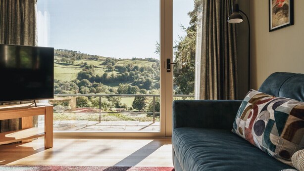 A living room with a television, a sofa, and a view of a green landscape through a glass door.