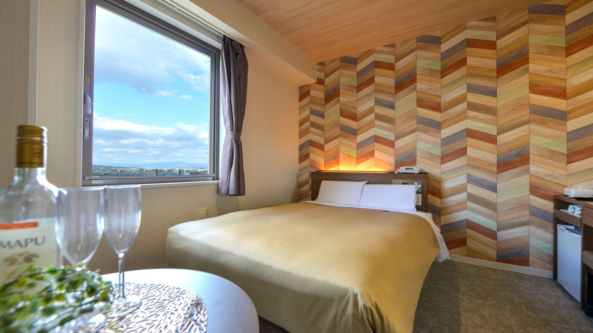 A hotel room with a bed, a nightstand, a window with a view, and a herringbone-patterned wall.