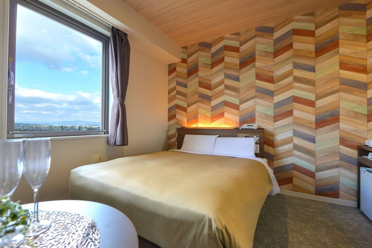 A hotel room with a bed, a nightstand, a window with a view, and a herringbone-patterned wall.