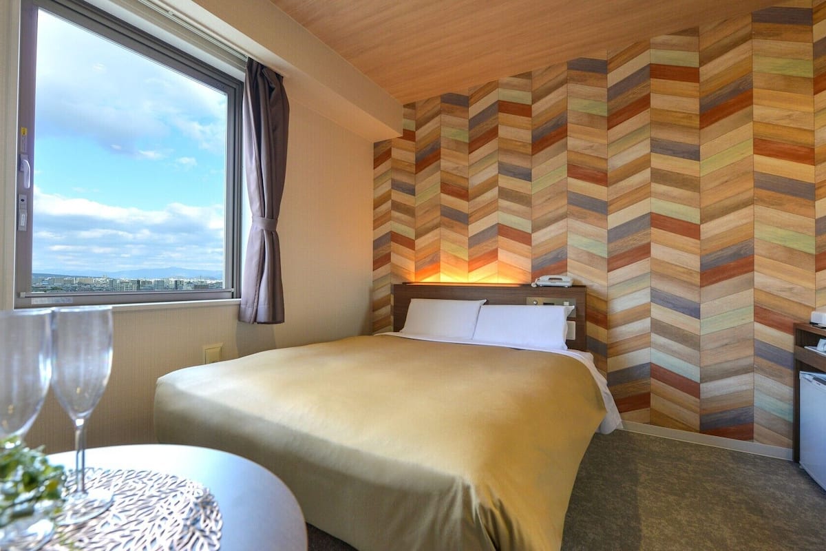 Habitación de hotel con cama, mesita de noche, ventana con vistas y pared con estampado tipo espiga.