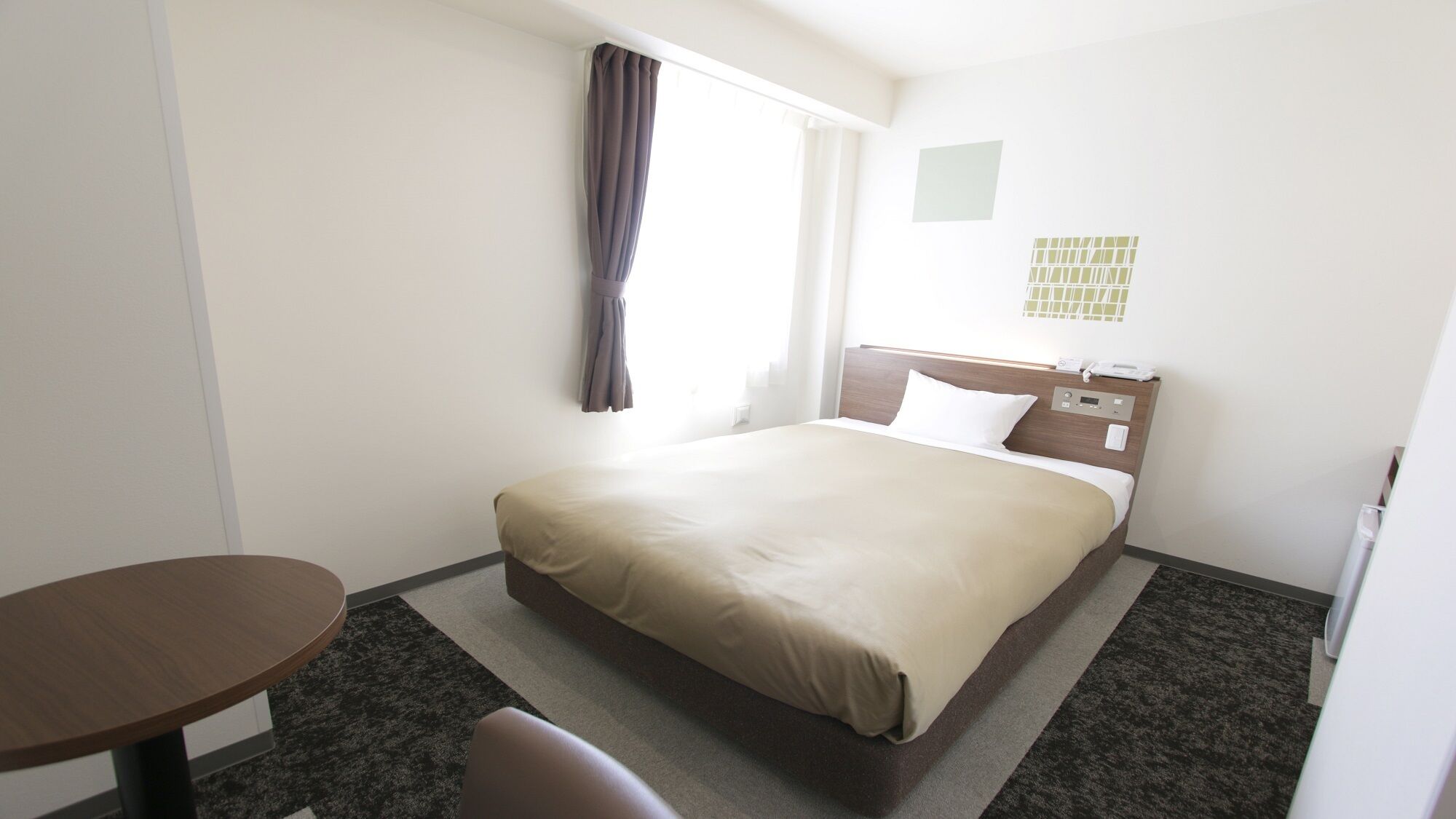 A hotel room with a bed, a desk, a chair, a window with curtains, and a wall-mounted phone.