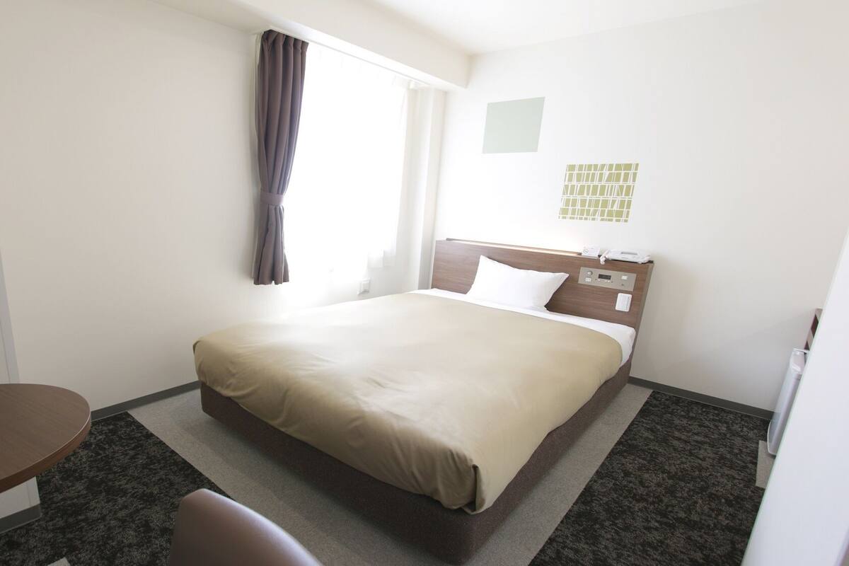 A hotel room with a bed, a desk, a chair, a window with curtains, and a wall-mounted phone.