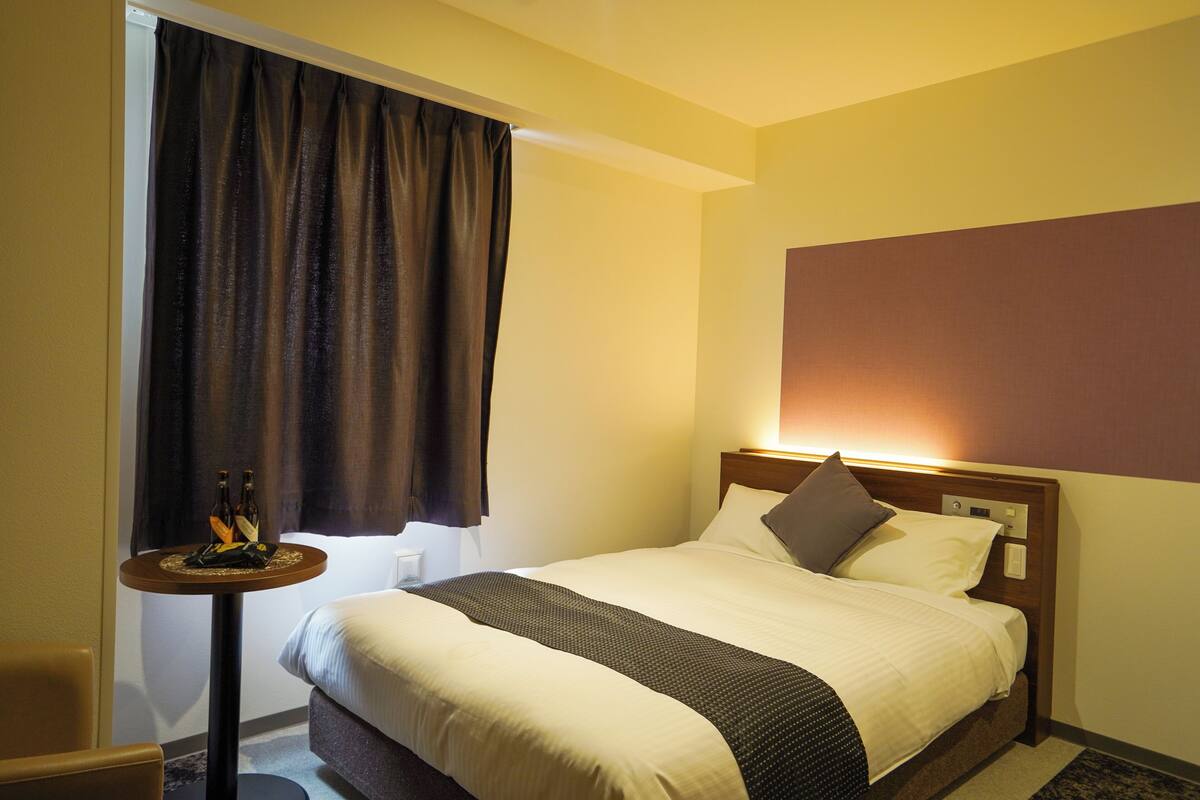 A hotel room with a neatly made bed, a small round table with a bottle and glasses, and a large window with curtains.