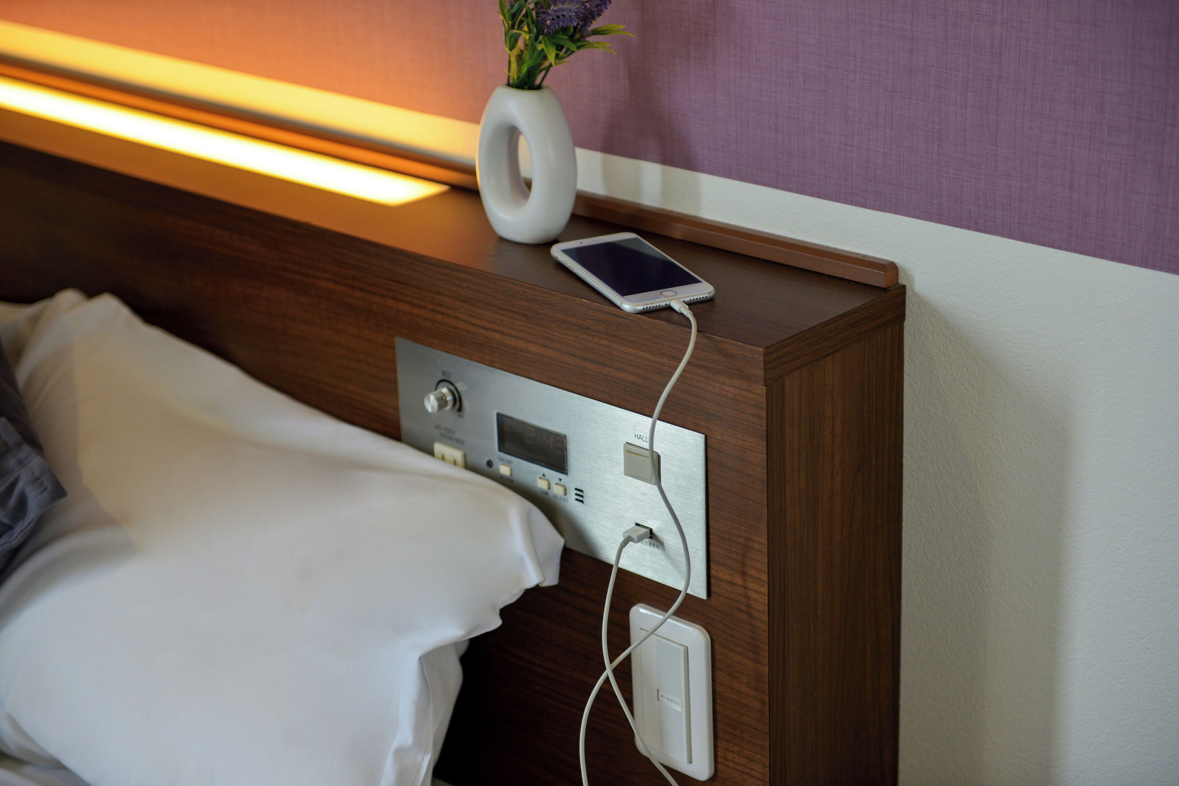 Ein Hotelzimmer mit einem Nachttisch aus Holz, einer weißen Vase mit Blumen, einem Smartphone und einer Bedienkonsole mit Steckdose.