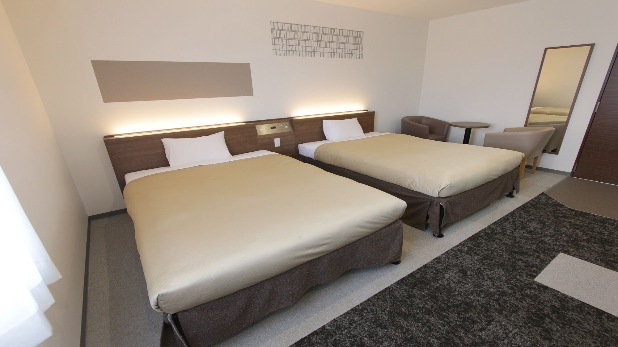 A hotel room with two beds, a small table, a chair, a mirror, and a wall-mounted light fixture.