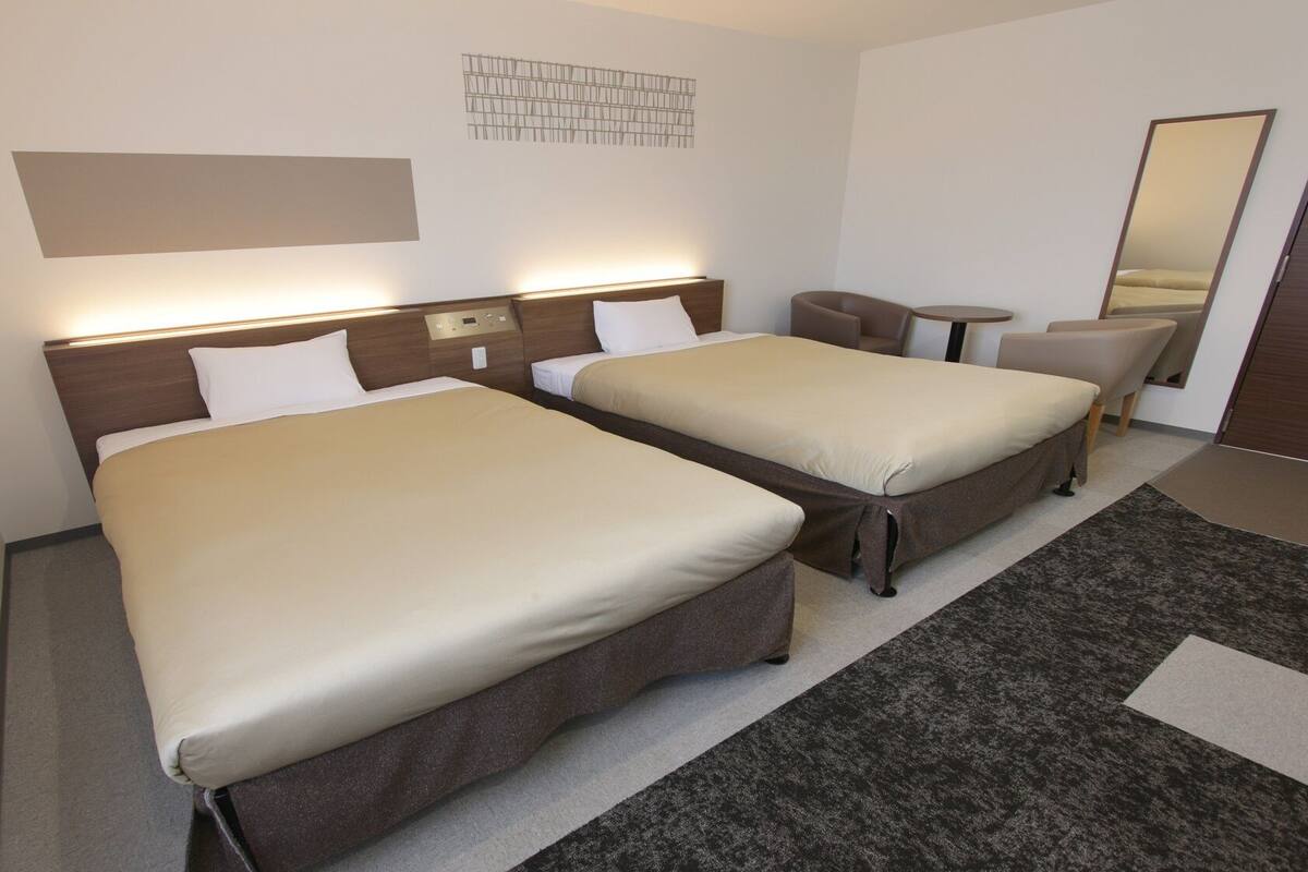 A hotel room with two beds, a small table, a chair, a mirror, and a wall-mounted light fixture.