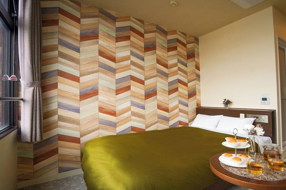 A hotel room with a bed, a side table with a tray of muffins and drinks, a wall with a chevron pattern, and a door.