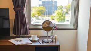 Escritorio de madera con un libro, una taza, un espejo y una ventana que ofrece vistas a edificios y árboles.